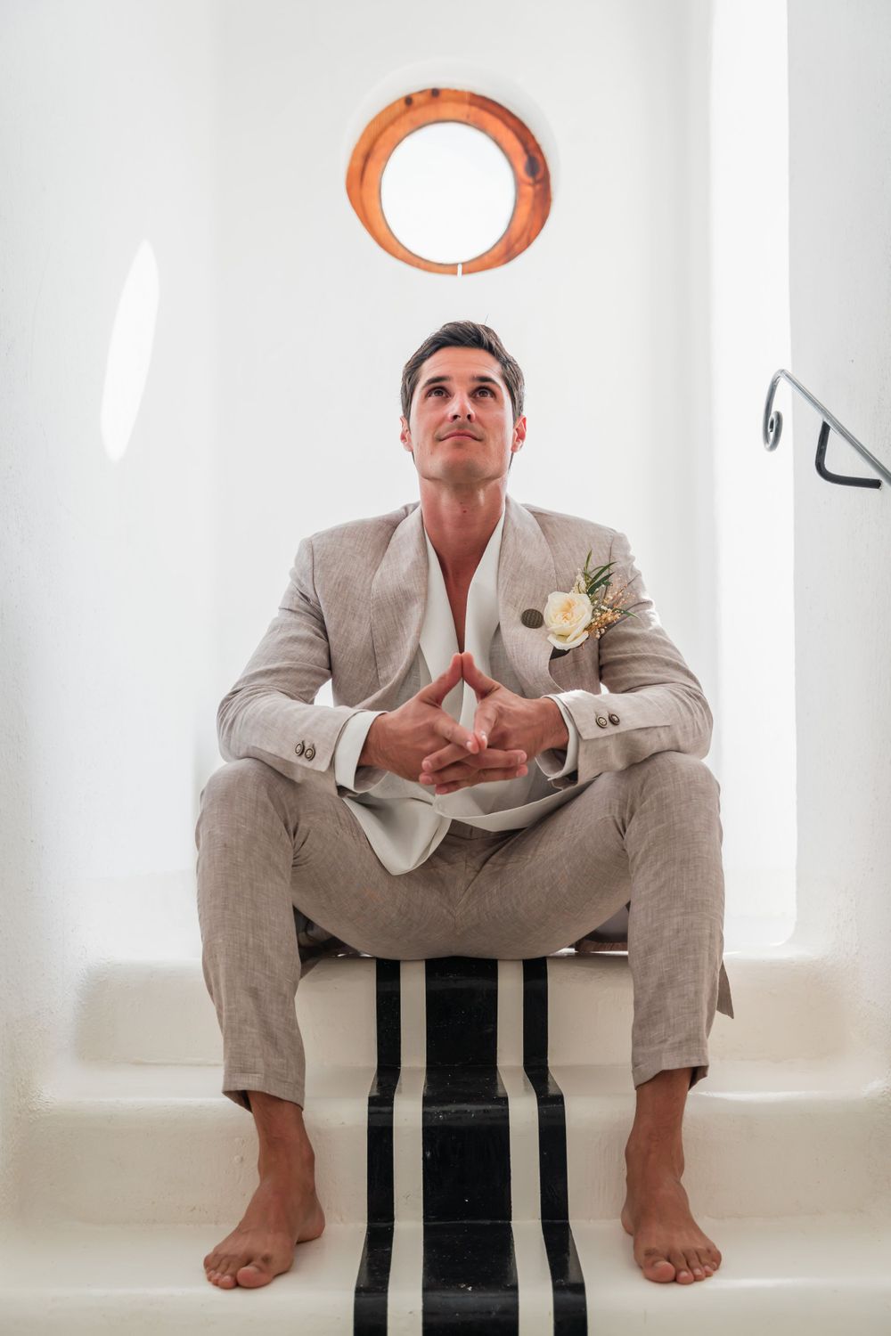 Wedding portrait featuring a light suited figure sitting barefoot with a boutonniere against a white wall with an orange porthole.