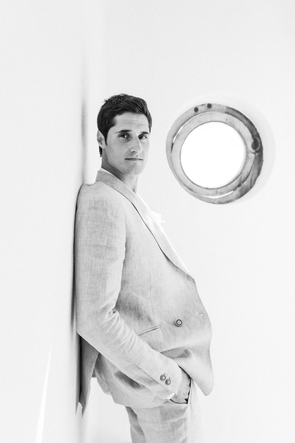 Black and white portrait of a person in a light linen suit standing against a white wall with a porthole.