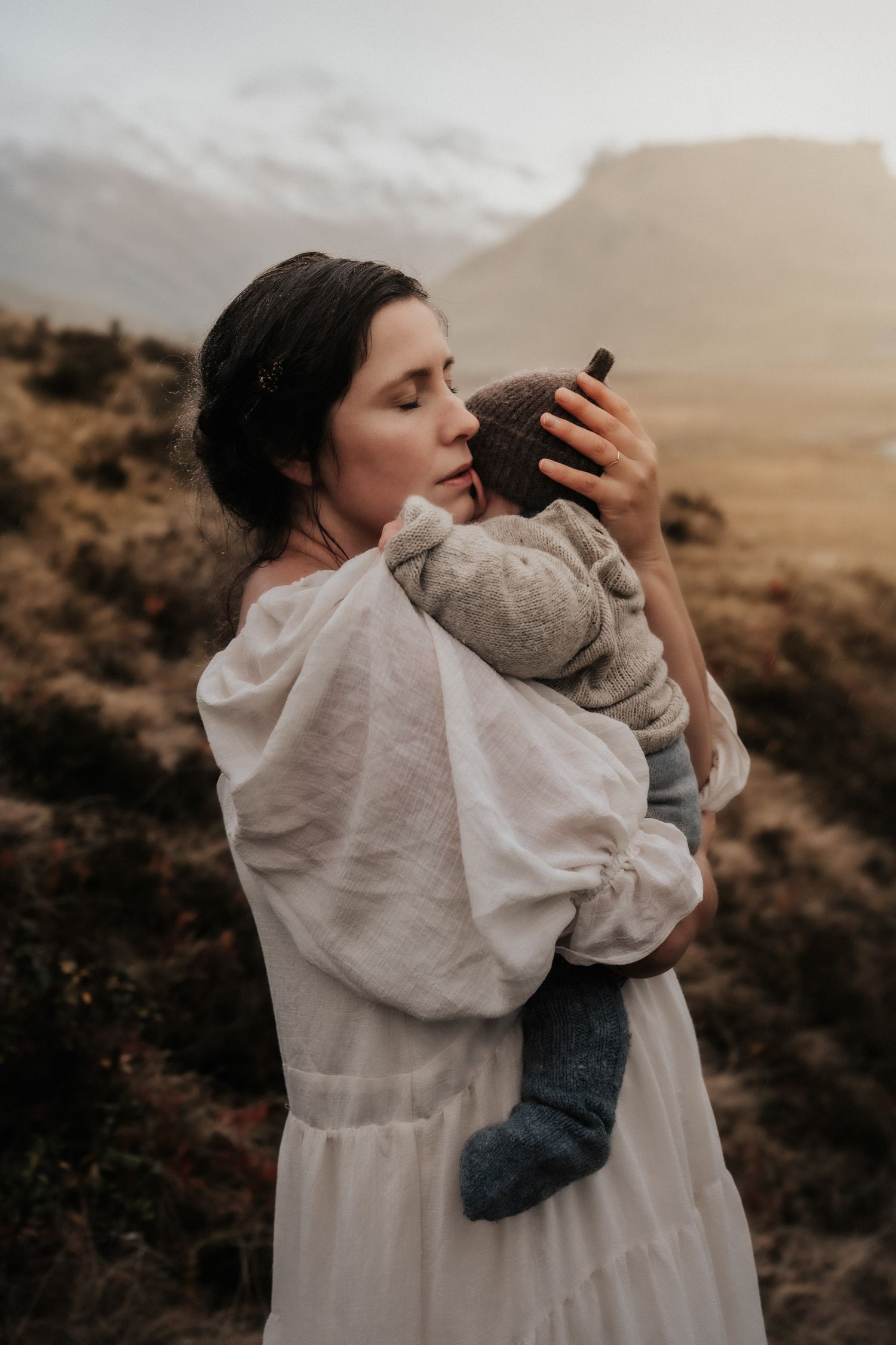 A tender moment between parent and infant against a mountainous landscape at dusk.