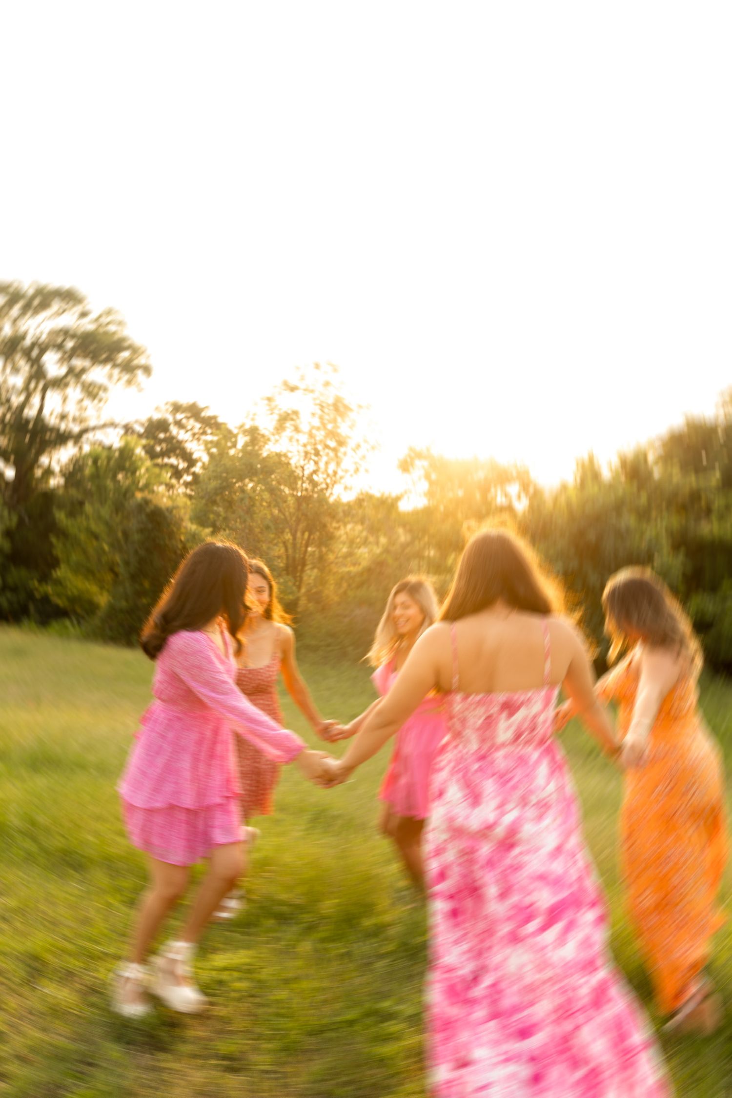 Motion-blurred photo of people in pink dresses dancing and spinning in a grassy field at sunset.