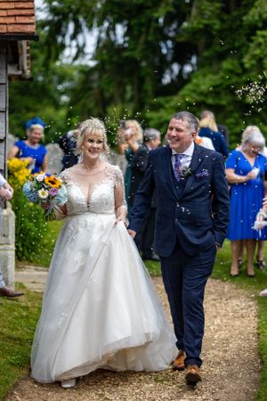 A wedding couple walks down a garden path while guests look on, with the bride carrying a sunflower bouquet.