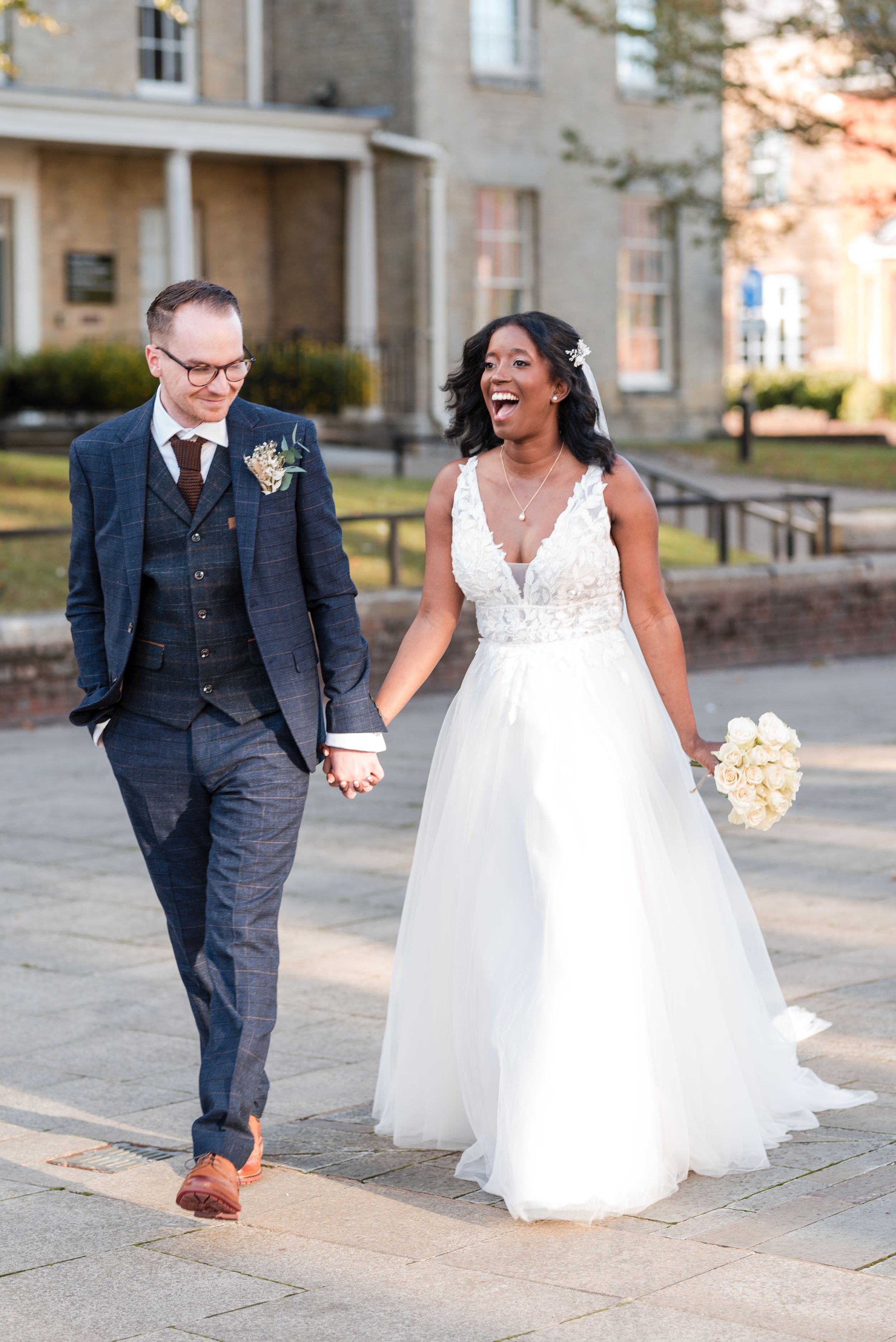A joyful newlywed couple walks together outdoors, the bride in a white gown and groom in navy suit.