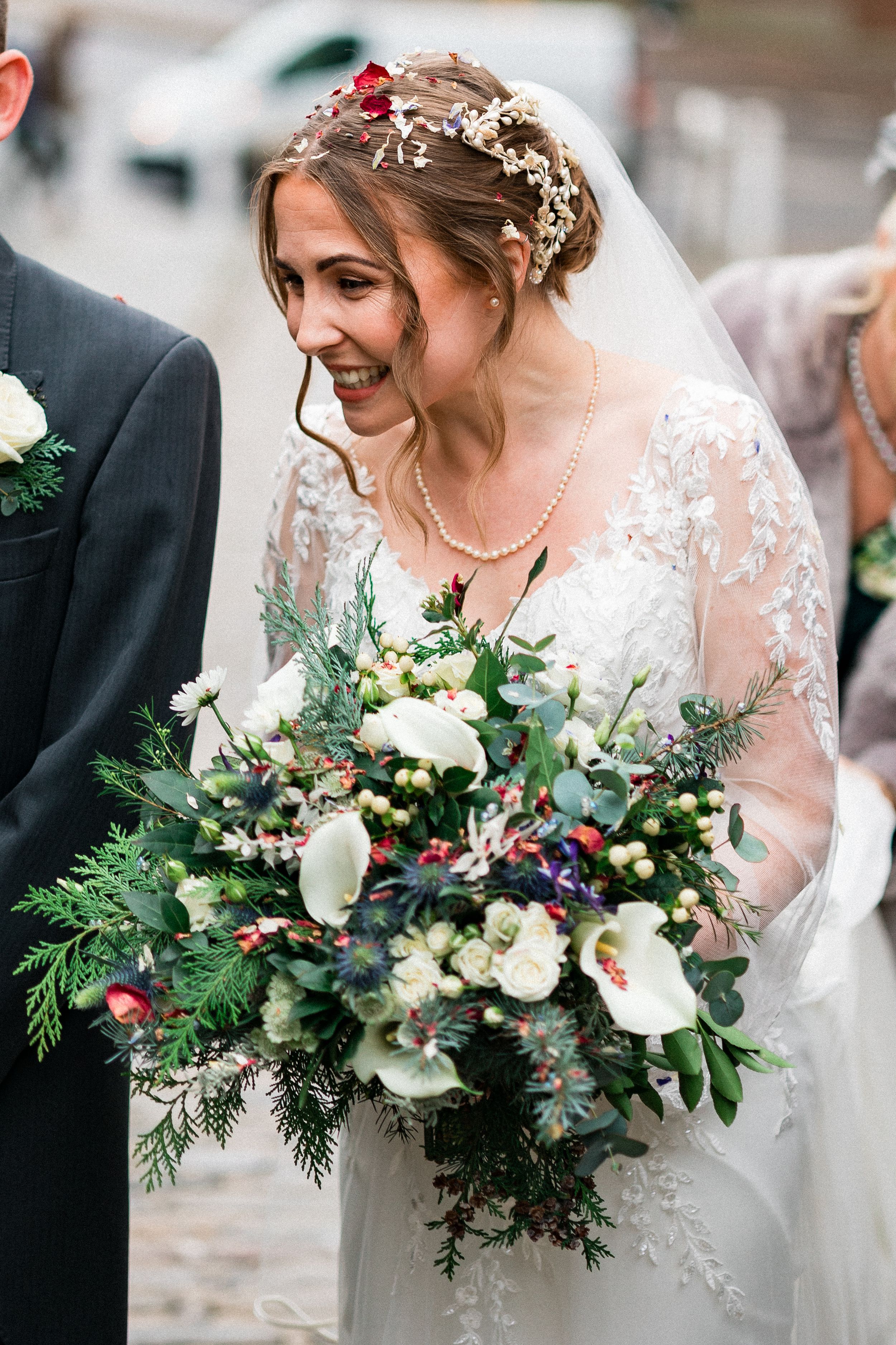 A bride holds a lush winter bouquet with white flowers and greenery while wearing a lace detailed dress.