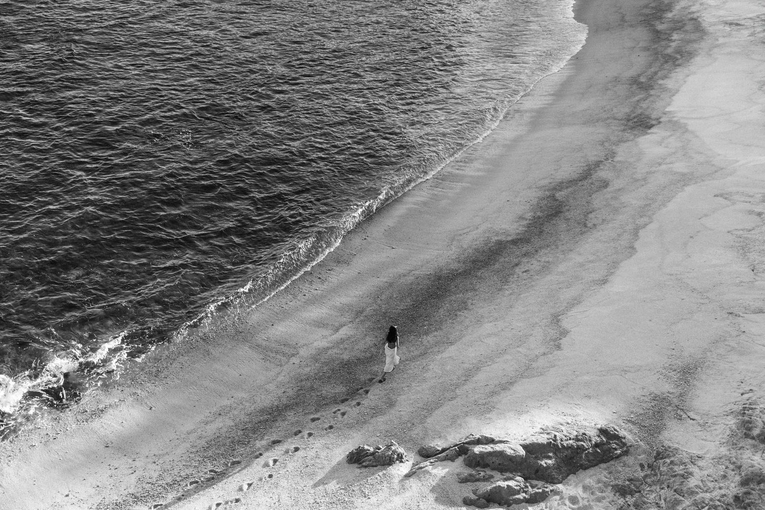 Aerial black and white view of lone figure walking along dramatic coastline.