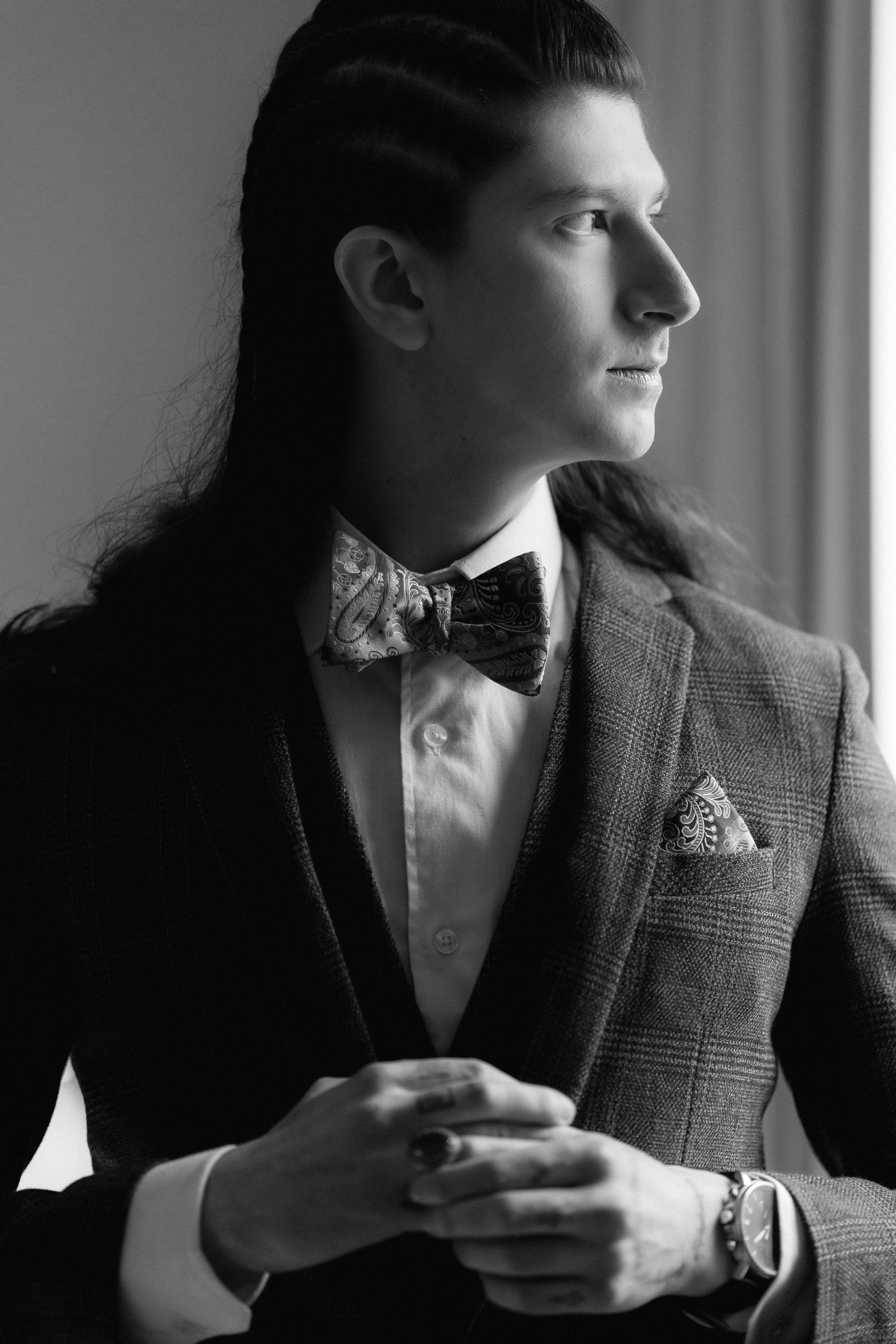 Person in a tweed jacket and bowtie sitting by a window in a moody black and white portrait series.