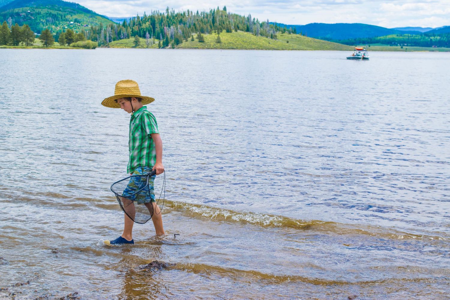 Someone strolling along a peaceful mountain lake shore wearing a sun hat.