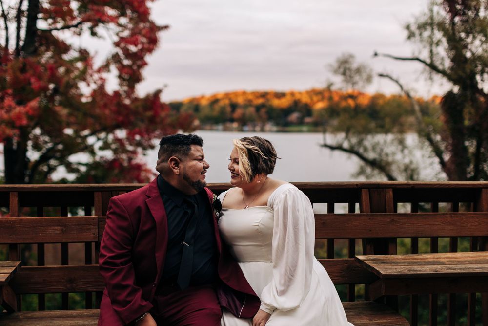 A romantic wedding couple share a joyful moment on a wooden deck overlooking an autumn lake landscape.