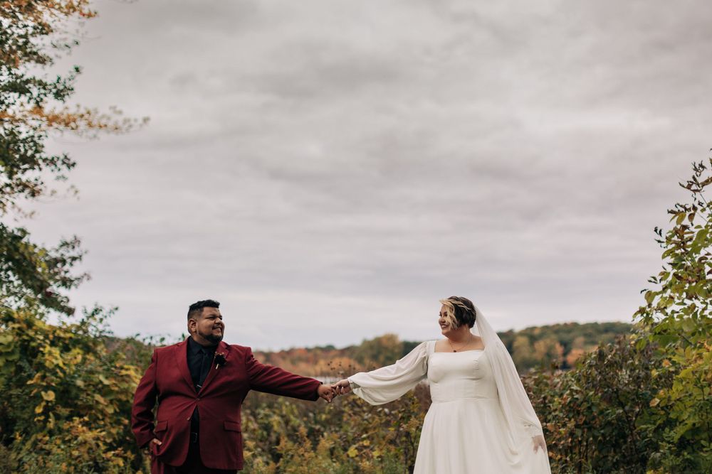 Wedding couple hold hands against a scenic fall backdrop with cloudy skies and colorful foliage.
