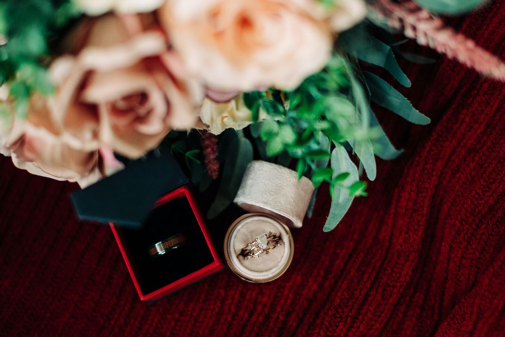 Wedding rings and floral decorations arranged on burgundy fabric with pink roses.