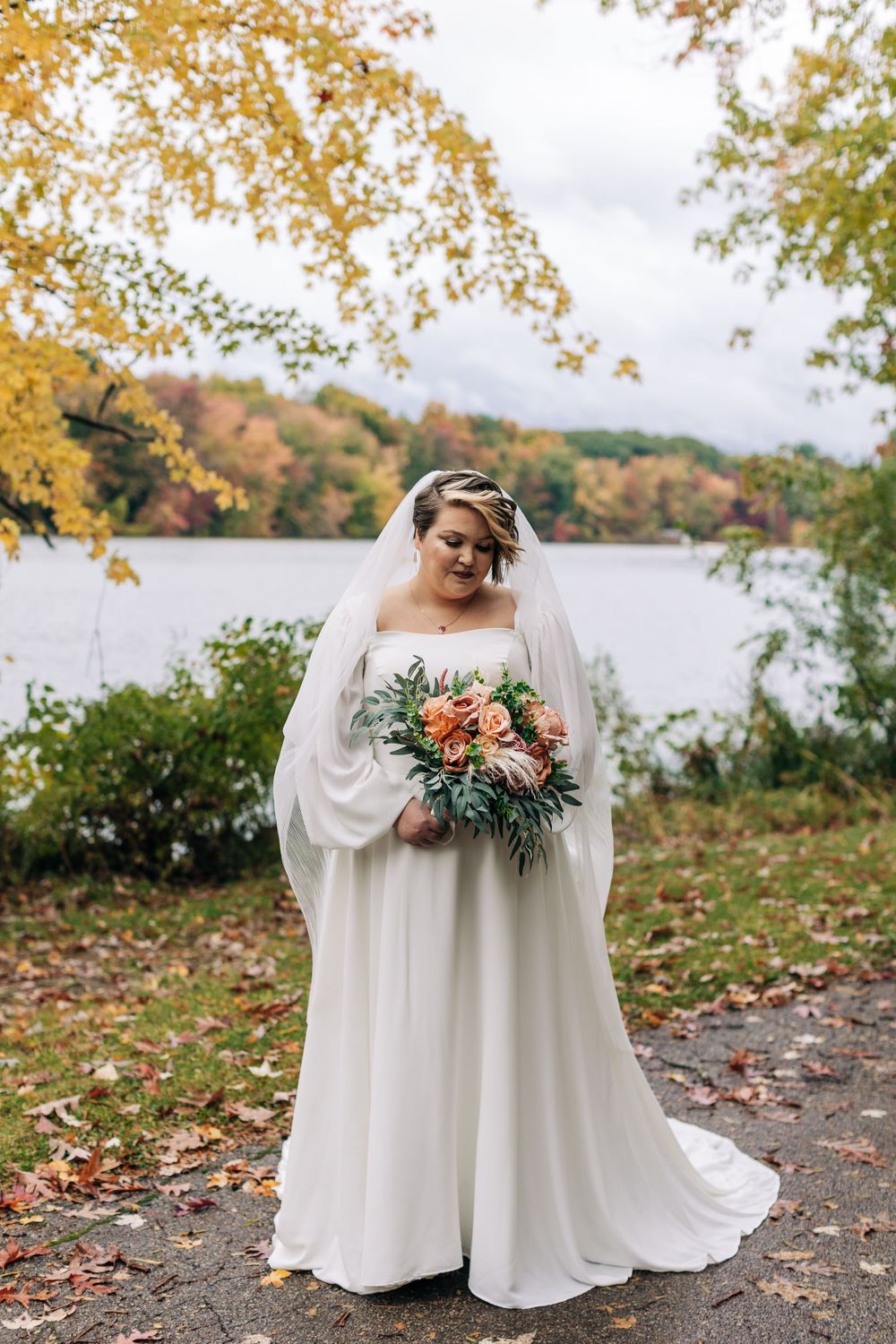 A bride in a flowing white dress holds a peach and green bouquet by a lake during autumn.