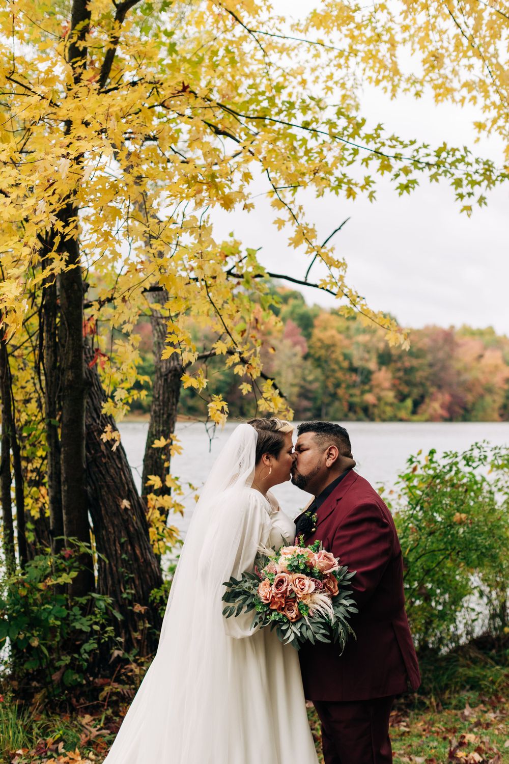 Newlyweds embrace underneath yellow fall foliage by a lakeside.