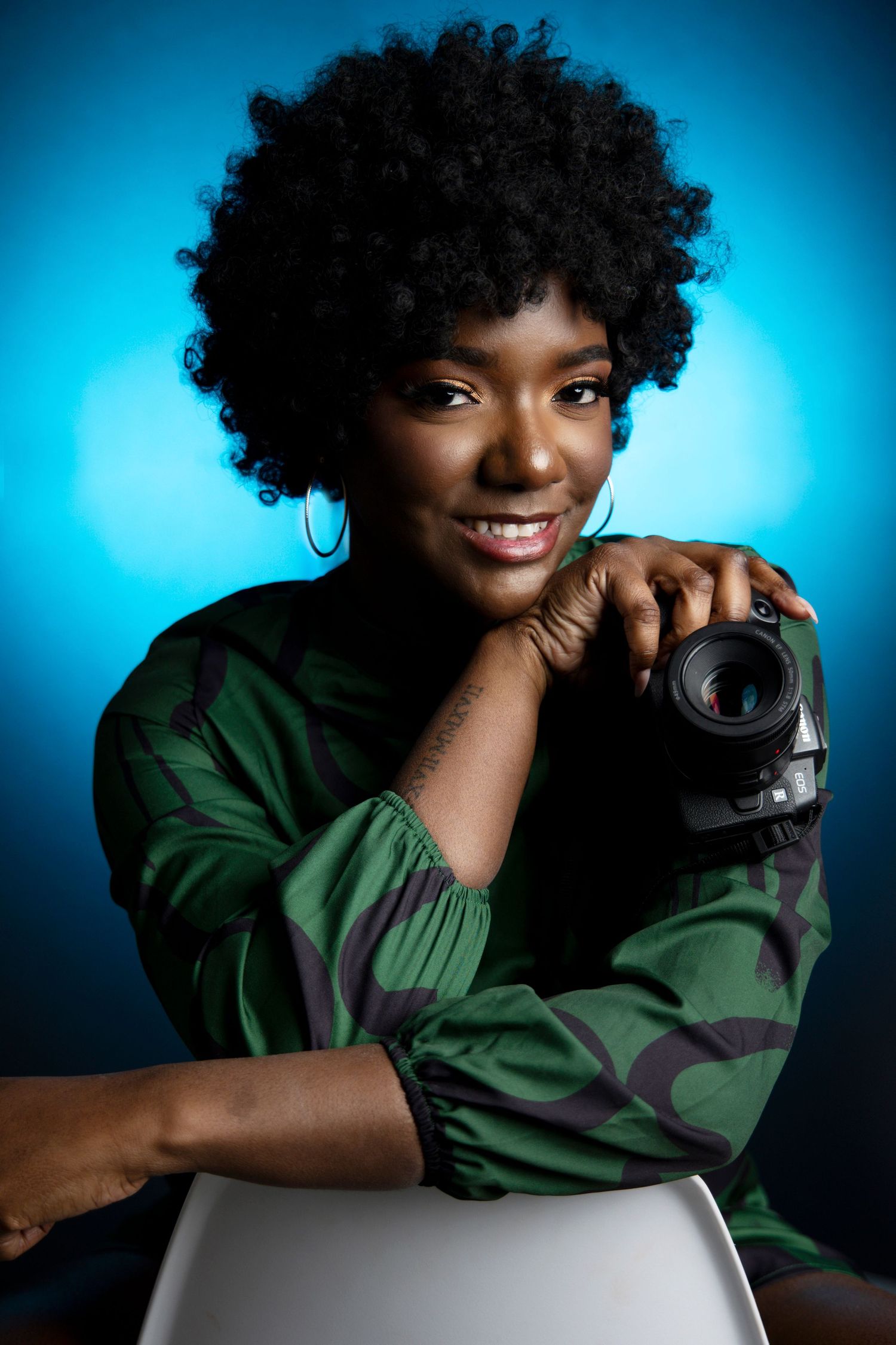 Professional photographer in green top holds camera against vibrant blue background.