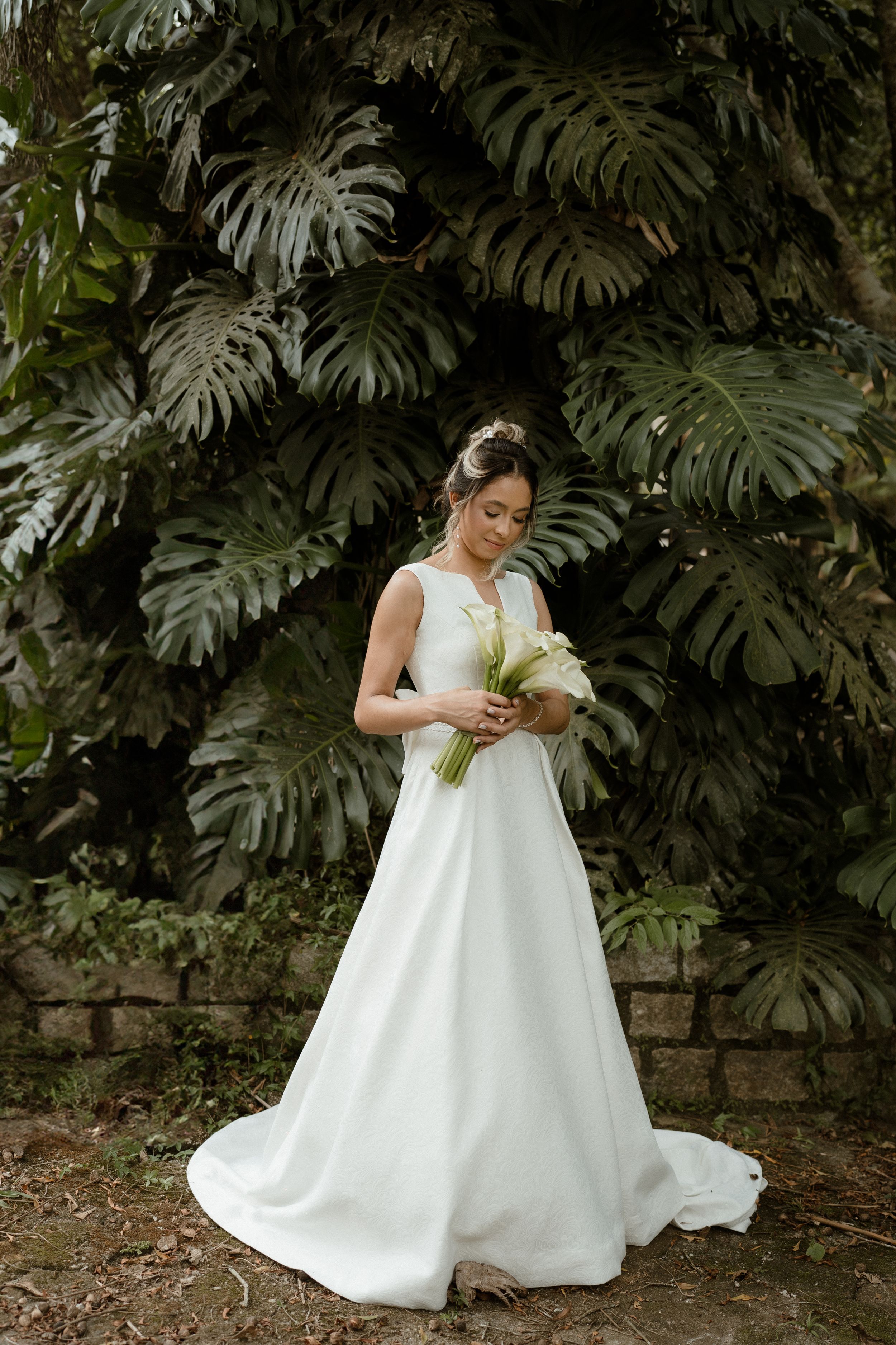 Bride in flowing white dress sits elegantly among tropical monstera leaves.
