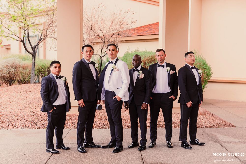 Groomsmen in black suits and ties stand in a line against a beige wall at an outdoor venue.