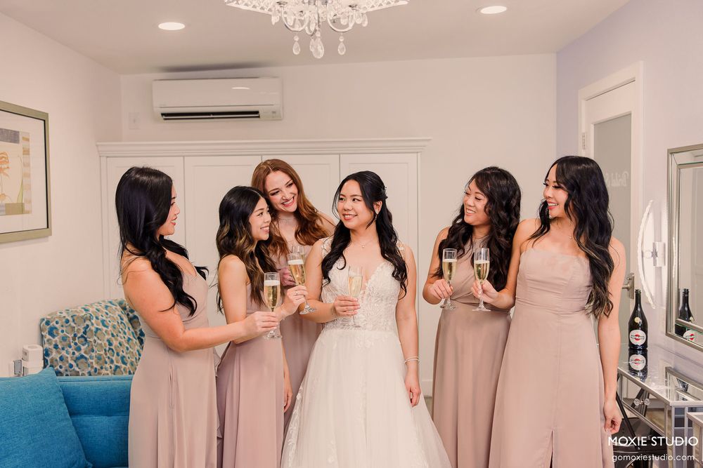 Bridesmaids in elegant champagne dresses gather in a bright room with a chandelier during wedding preparations.