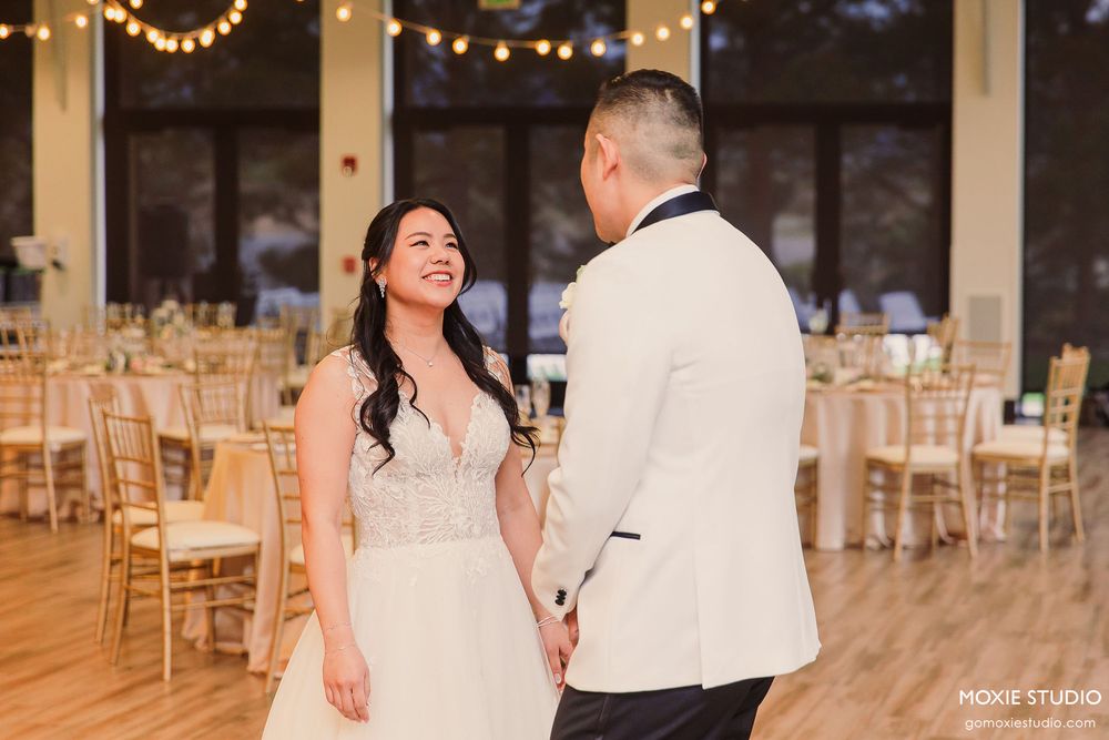 A couple shares a joyful moment in an elegant reception hall decorated with string lights and wooden chairs.