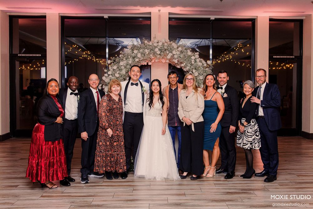 Full wedding party and family formal portrait in ballroom with pink lighting and floral decorations.