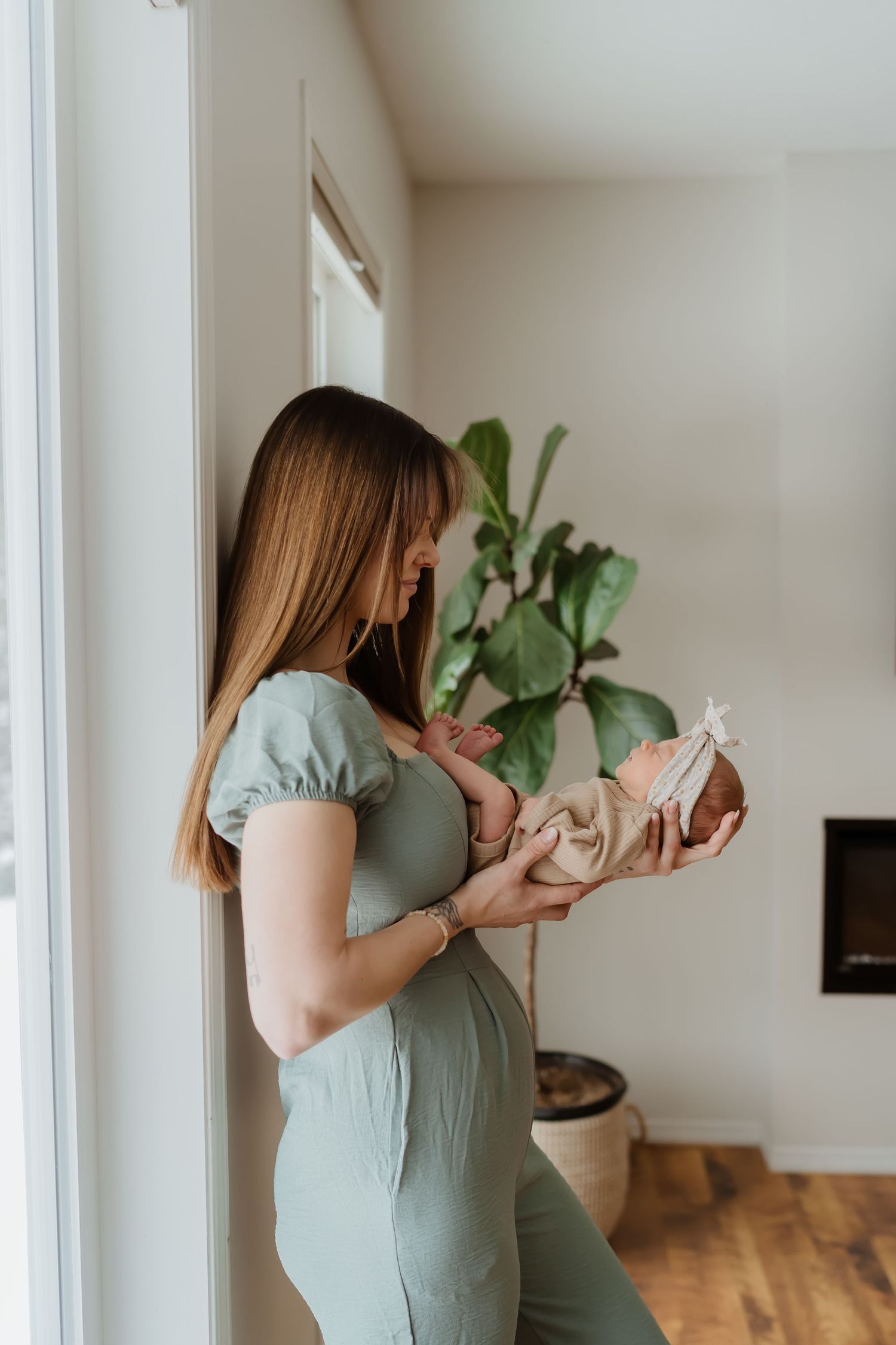 Woman in mint dress holds tiny baby shoes while standing by window with indoor plant.
