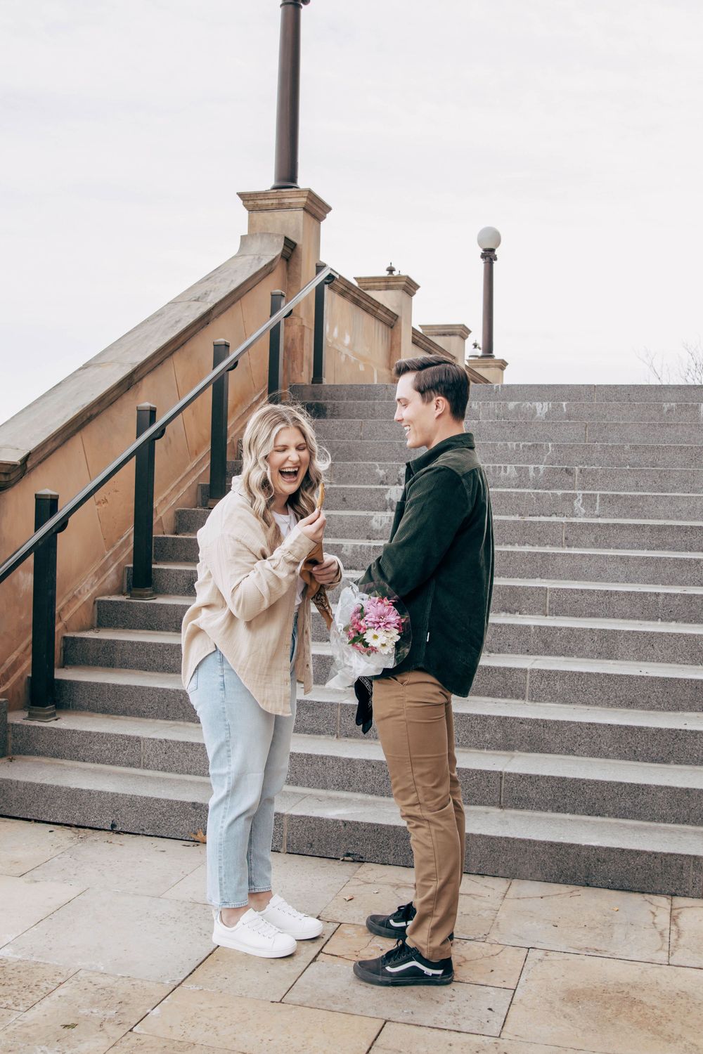 A couple sharing a joyful moment on stone steps with one holding a pink bouquet.