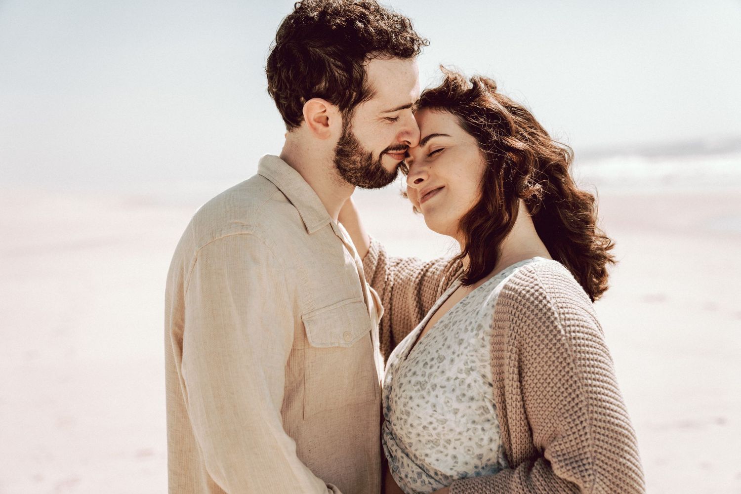 Romantic couple sharing an intimate moment on a beach, touching foreheads and smiling softly.