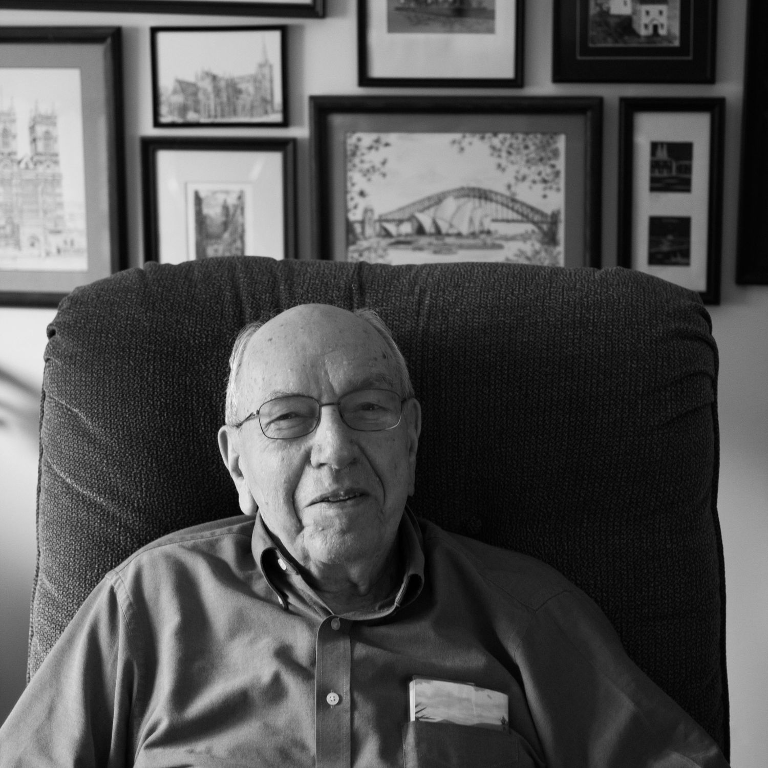 elderly man smiles at camera, behind him are pictures from his travels