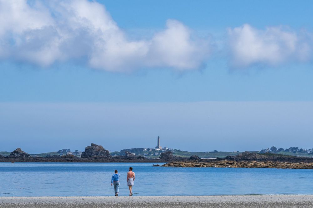 Two people walking on beach with rocky coastline and lighthouse in background.