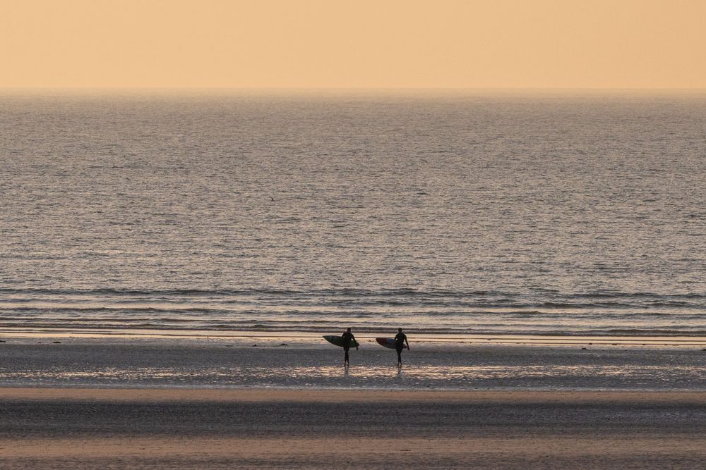 Two silhouetted figures standing on beach at sunset with orange sky.