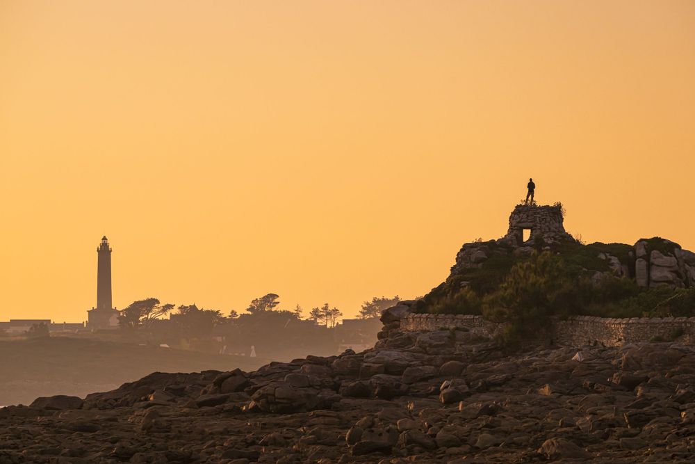 Silhouetted lighthouse and church on rocky coastline at golden sunset.