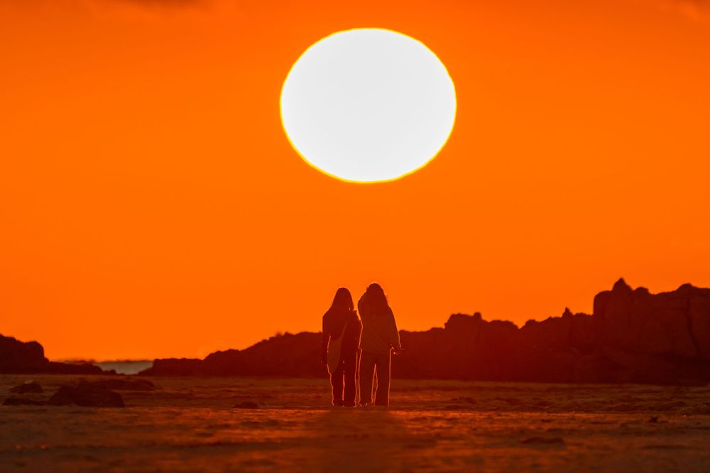 Silhouetted figures against large orange sun setting over mountain landscape.