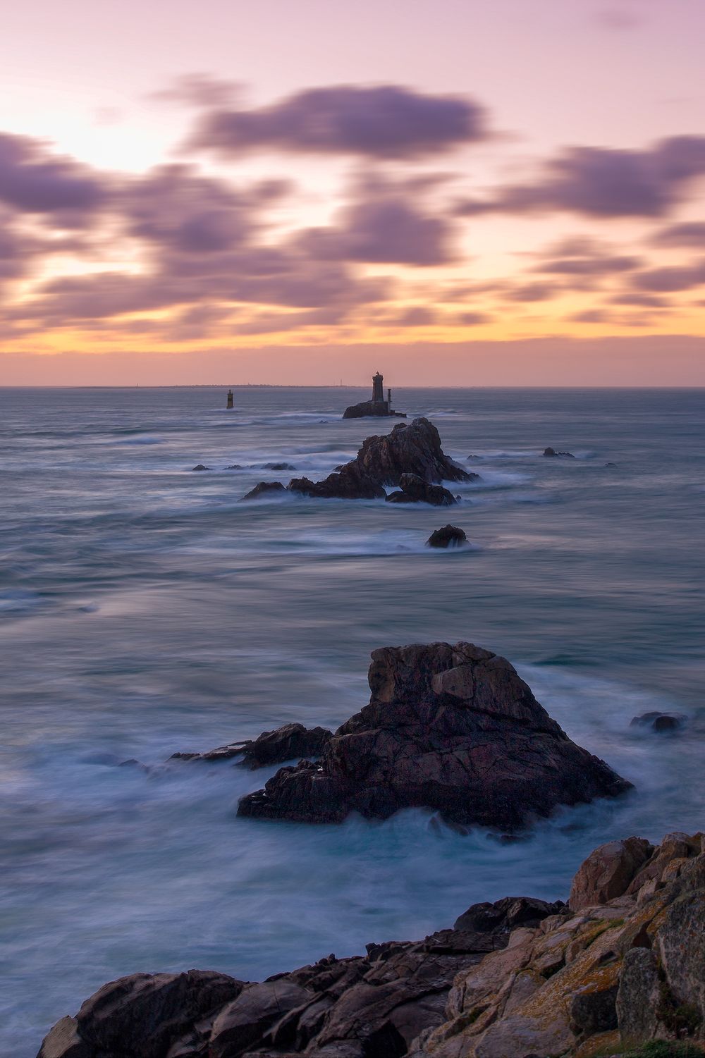 Dramatic sunset over rocky coastline with lighthouse silhouette and crashing waves.