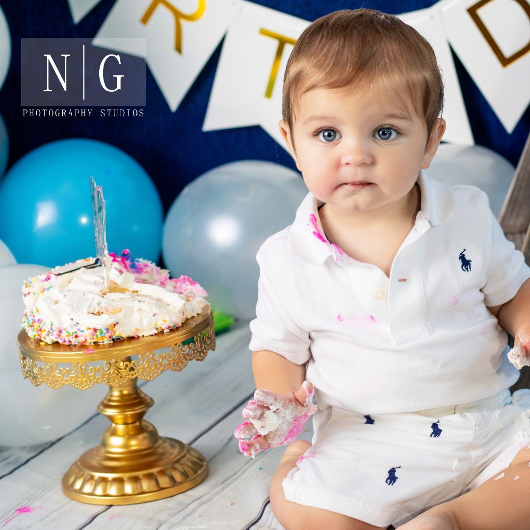 First birthday cake smash setup with blue balloons and gold cake stand against navy backdrop.