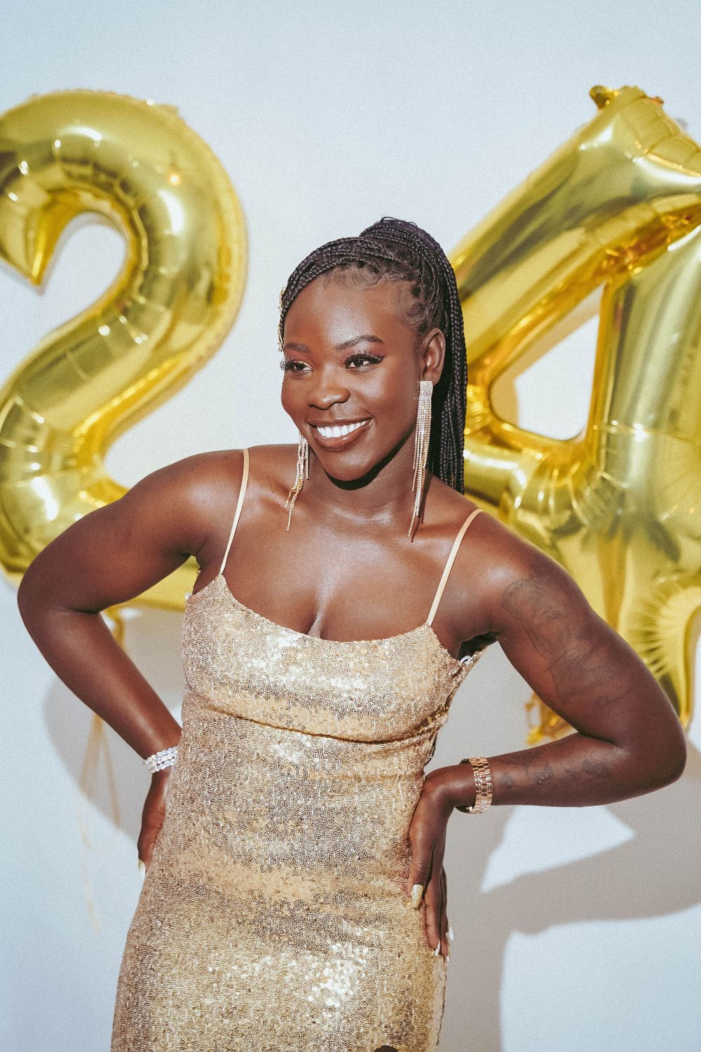 Model posing in front of gold balloon numbers wearing sparkly gold dress for celebration photos.