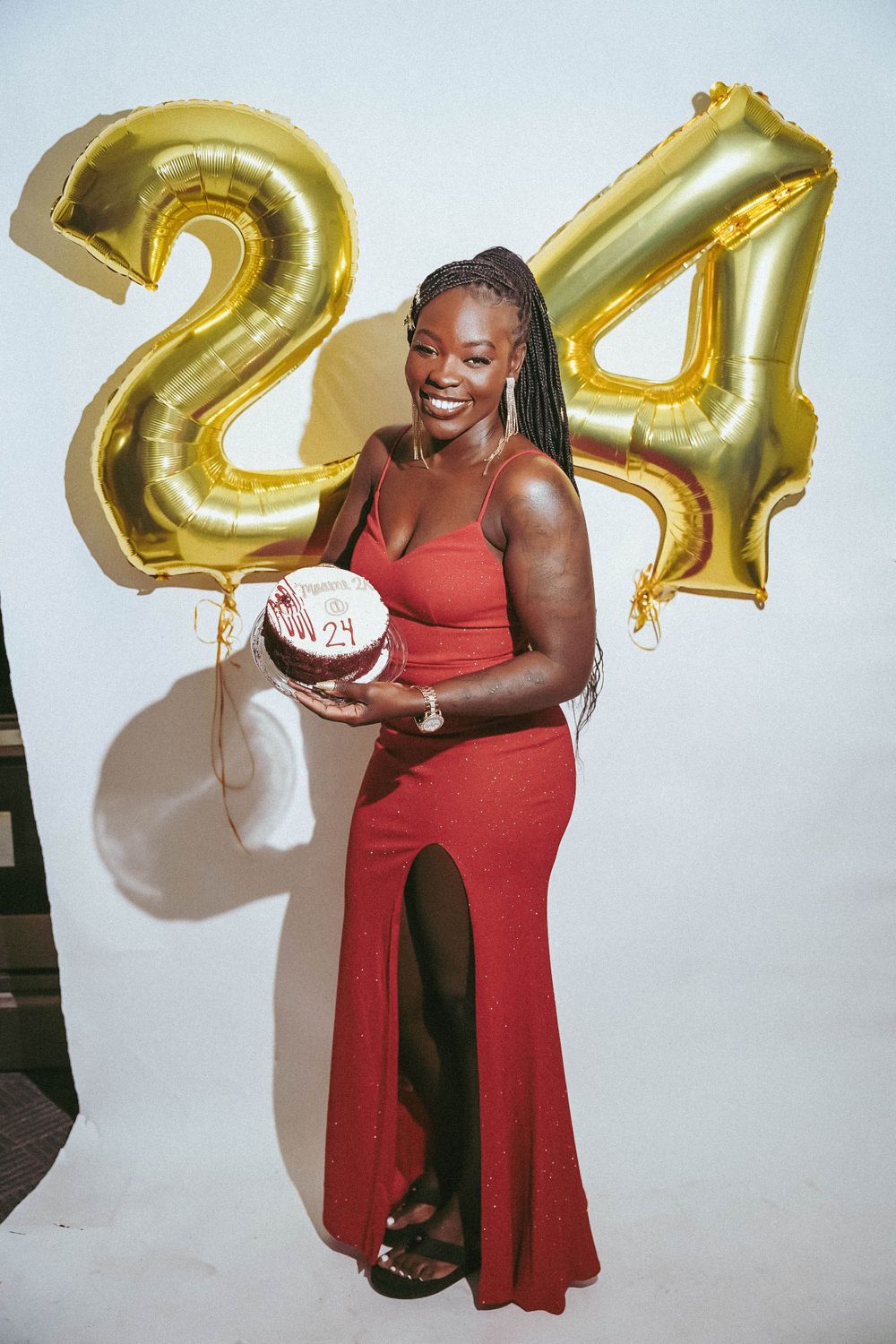 Woman in red dress poses with birthday cake in front of gold number balloons.