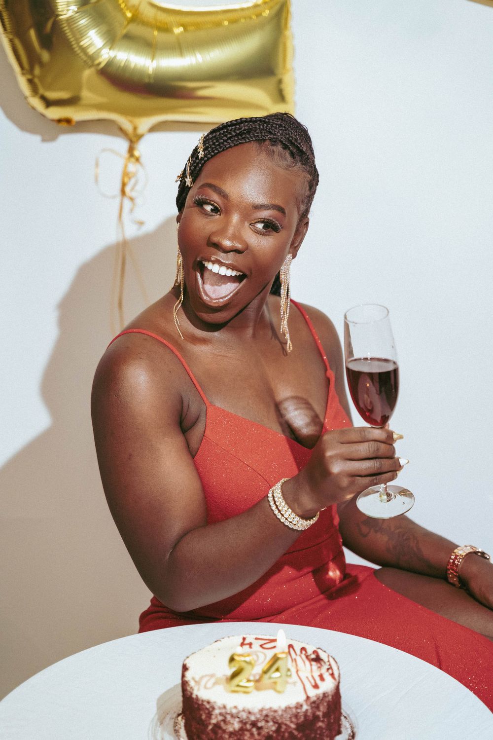 Woman in red dress celebrates with cake and wine against backdrop with gold balloons.