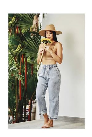 Woman in tan bralette jeans and wide brim hat holding sunflower near tropical palm plants.