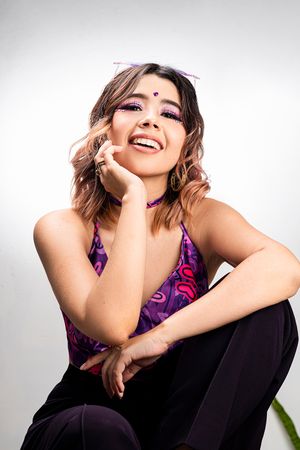 Smiling young woman with purple makeup and colorful top sitting casually against a white background.