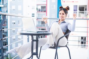Smiling businesswoman relaxing on balcony with laptop raising glass in urban setting.