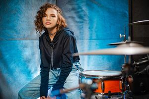 Young woman with curly hair sitting at drum kit in music studio with blue backdrop.