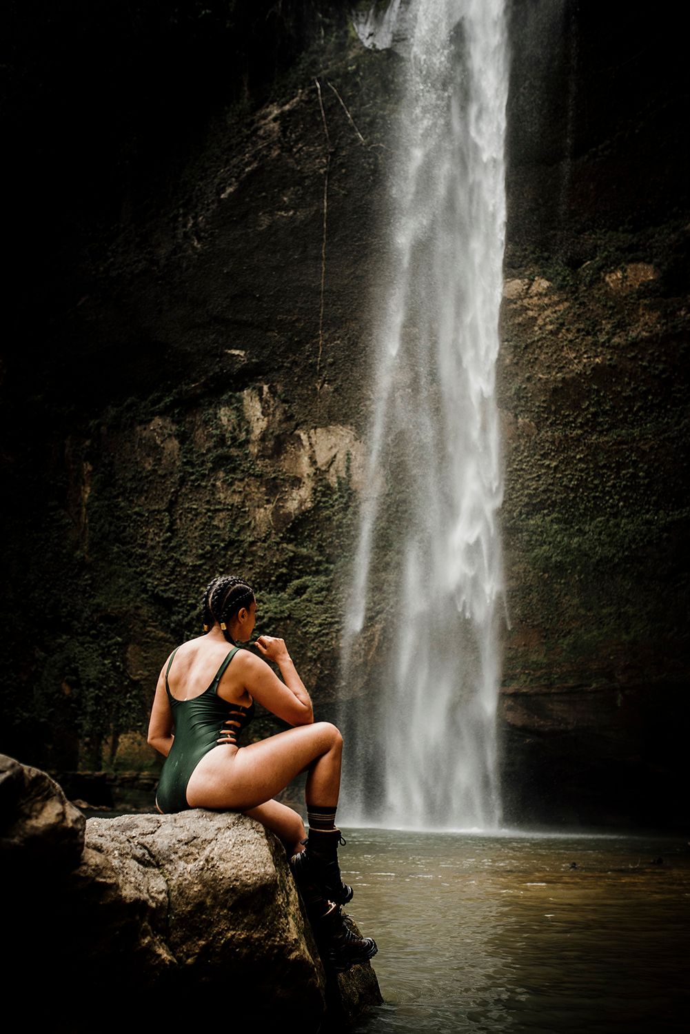Woman in green swimsuit sitting on rock admiring tall waterfall in dark cave setting.
