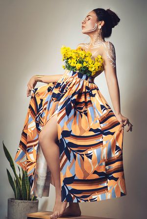 Woman in colorful tropical print maxi dress holding yellow flowers with eyes closed on stool.
