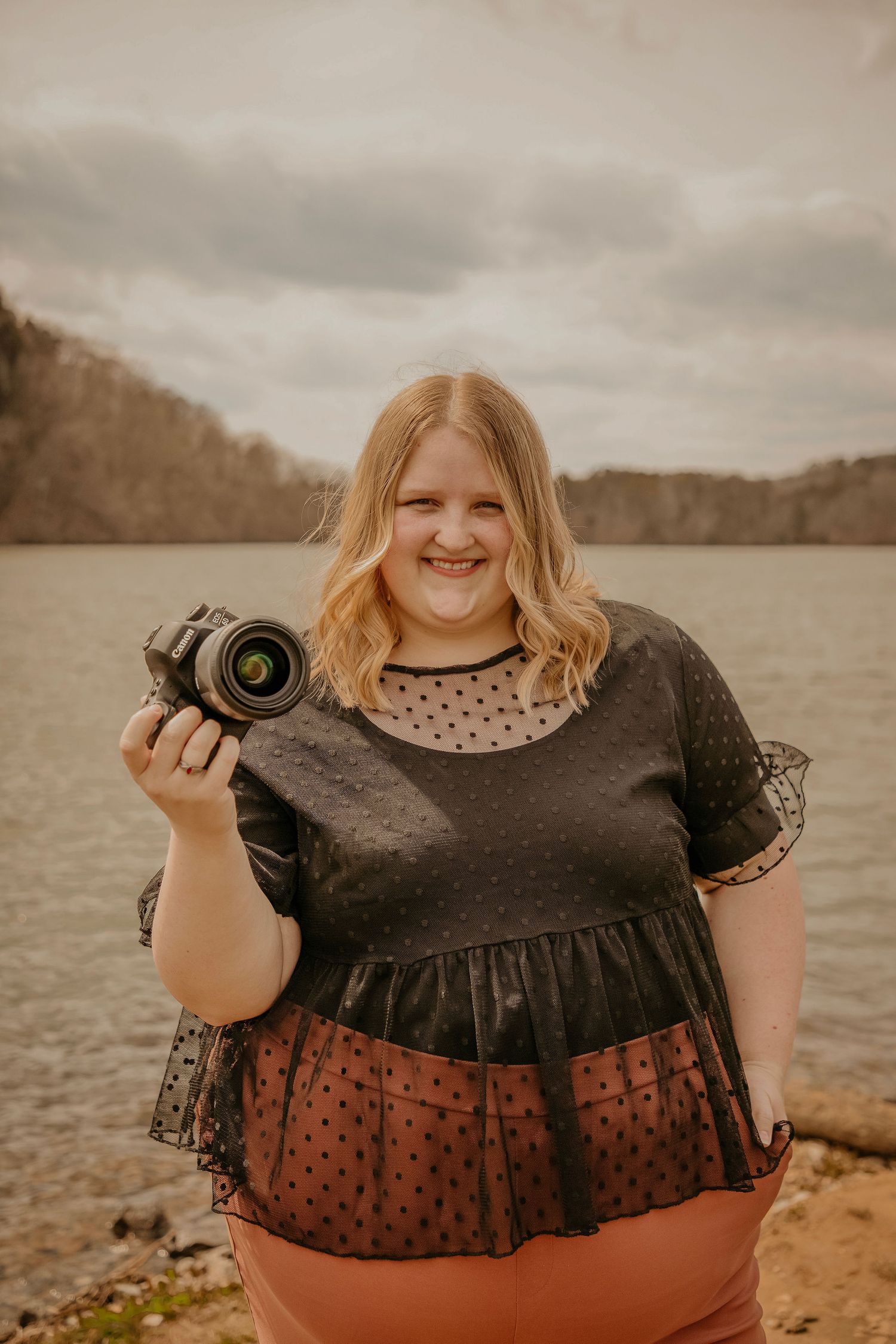 Young photographer in a black lace top holds a DSLR camera while standing near a lake on an overcast day.