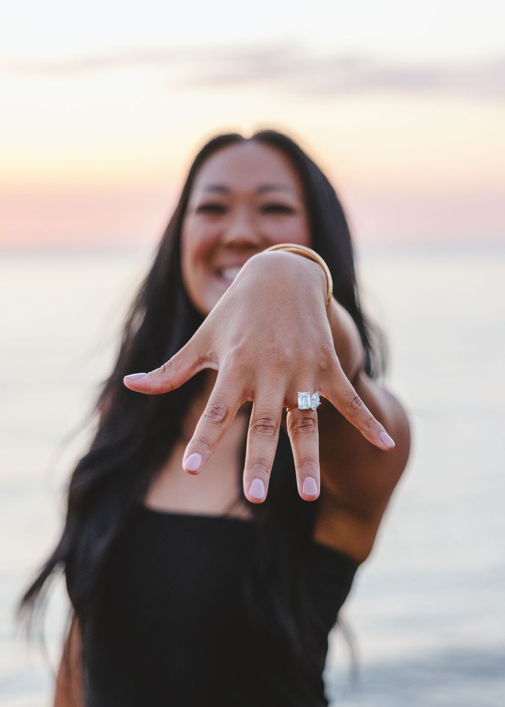 Engagement ring sparkles on hand held up at sunset against a blurry beach backdrop.