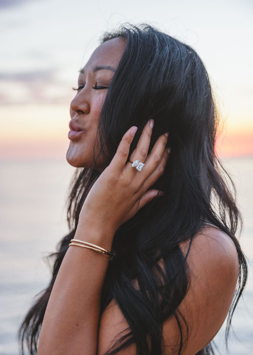 Profile silhouette of a woman with long dark hair and engagement ring posing at sunset by the ocean.