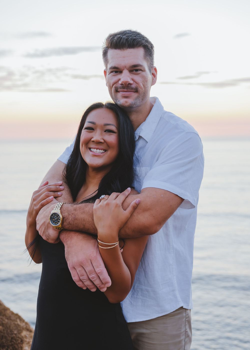 Couple embraces during sunset at beach, wearing casual white and black outfits against ocean backdrop.