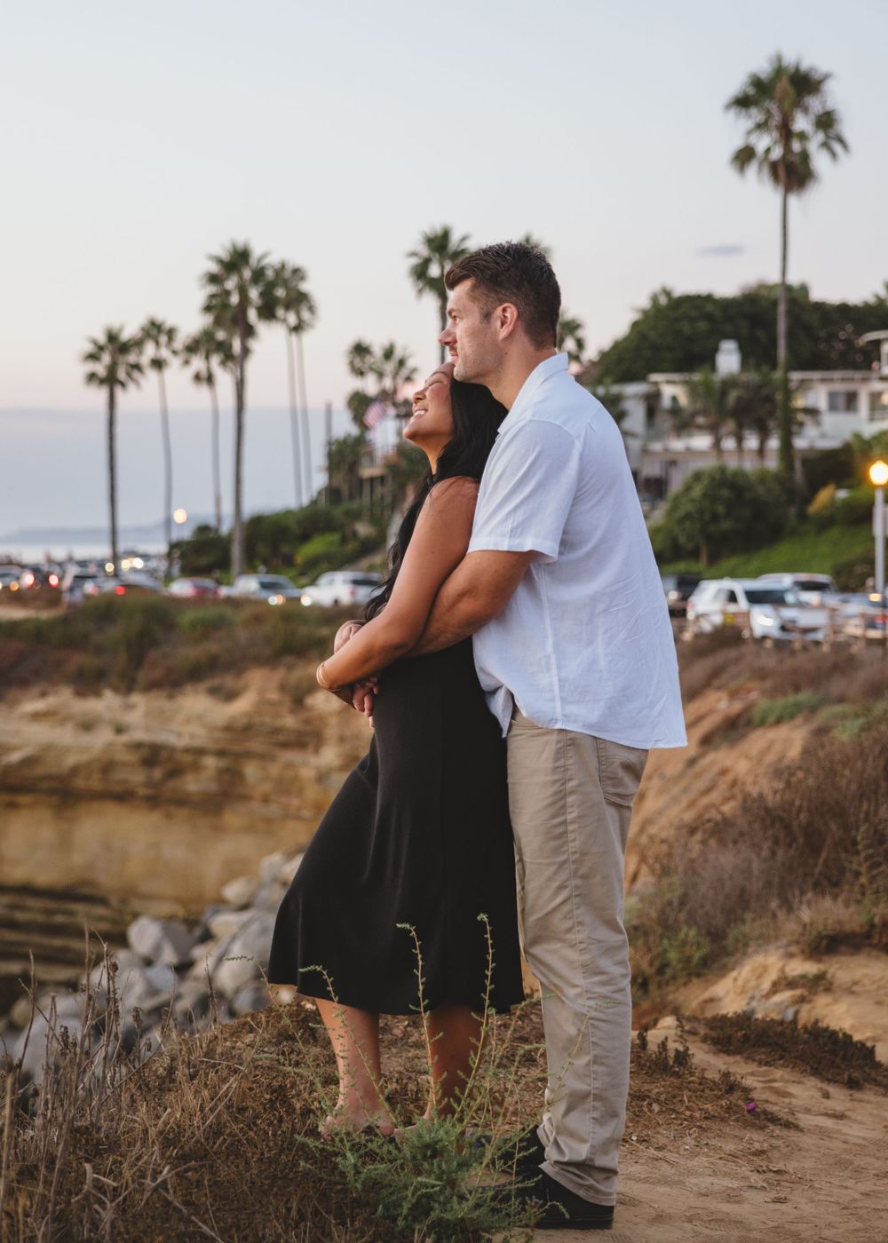 A couple embraces while standing on a coastal cliff with palm trees and ocean views in Southern California at sunset.