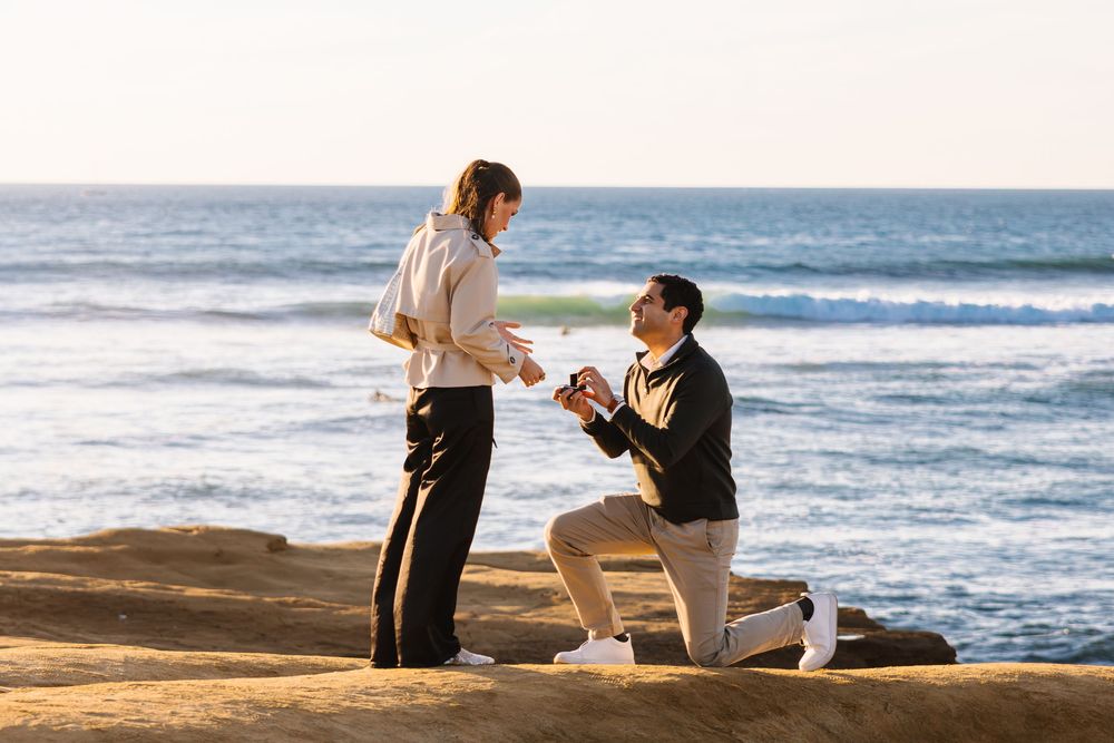 A couple shares a romantic proposal moment on a rocky beach at sunset with ocean waves in the background.