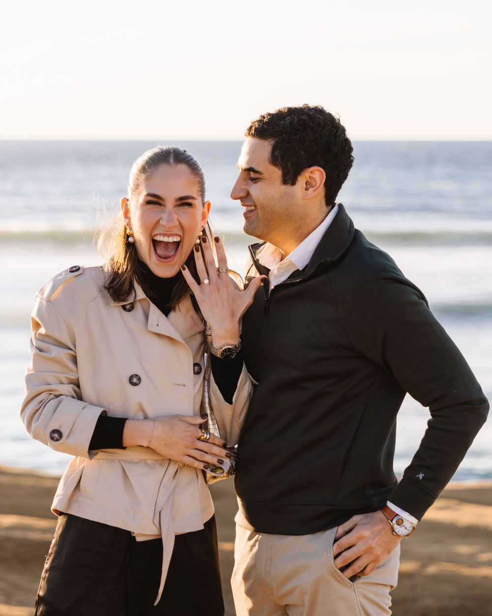 A joyful couple shares a romantic moment on the beach at sunset, laughing and embracing in casual attire.