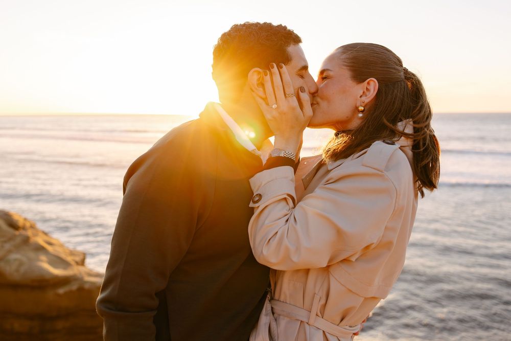 A couple shares a romantic kiss on the beach at sunset with golden light illuminating the scene.