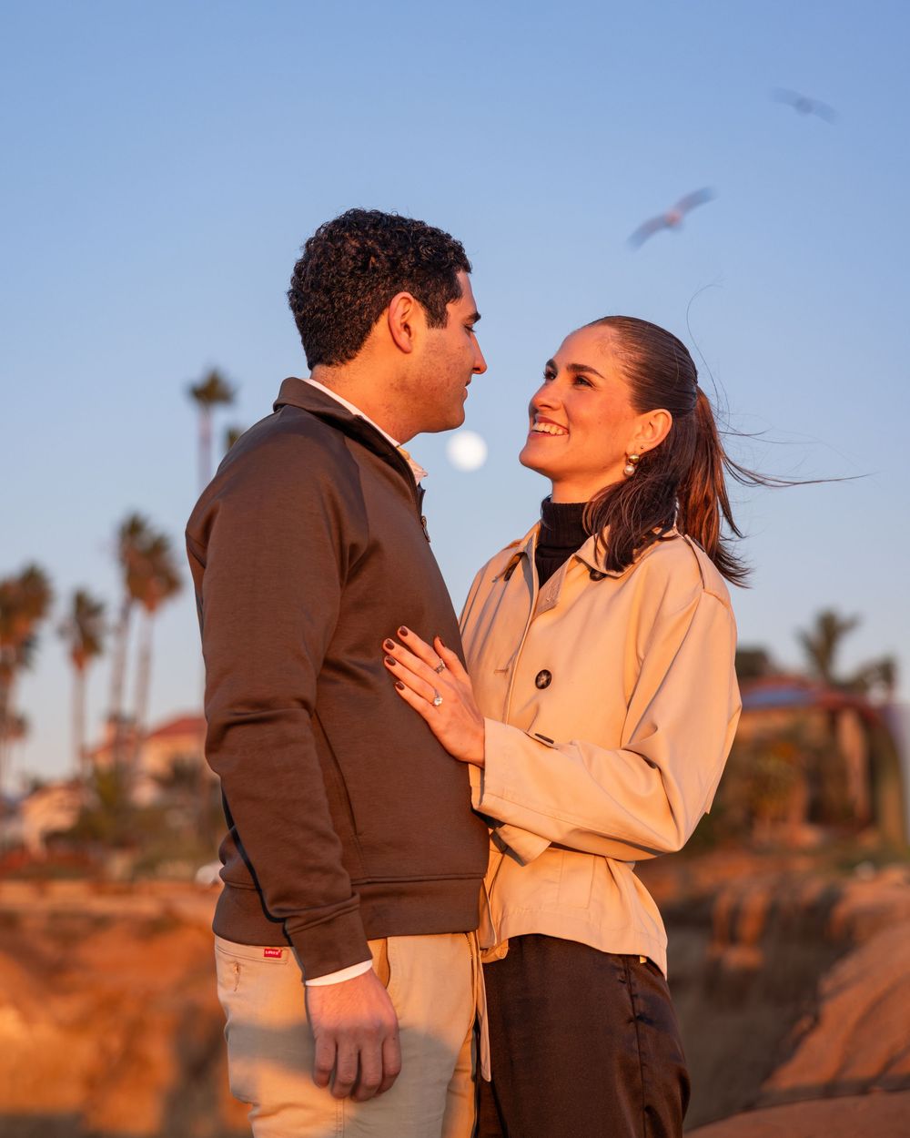 A couple shares a joyful moment together against a backdrop of palm trees and blue sky at sunset.
