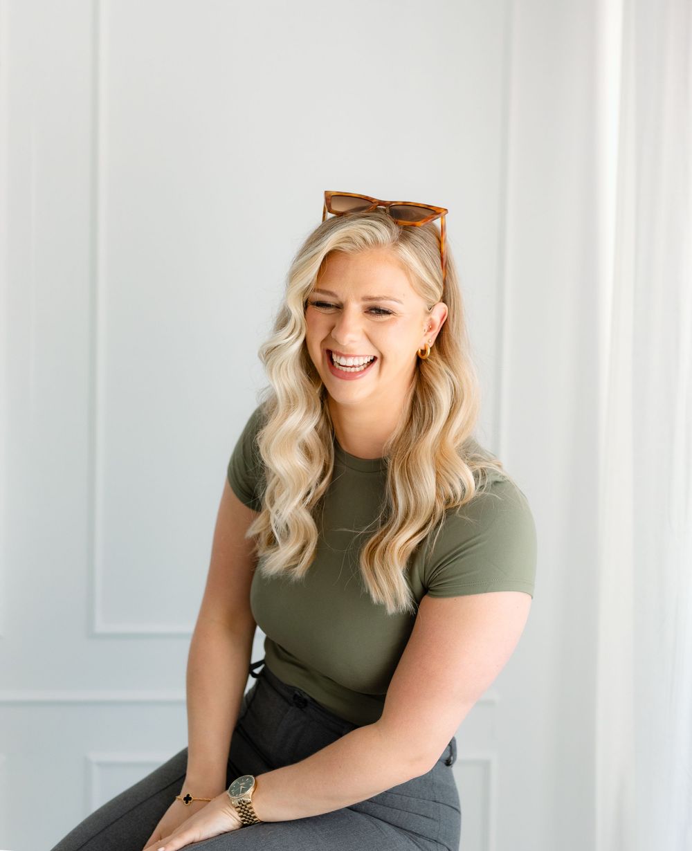 Person in olive green top sitting casually against white paneled wall in bright indoor setting.
